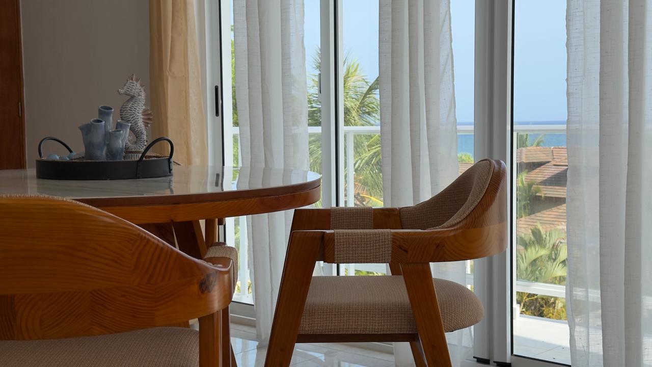 Dining area with wooden chairs, glass table, and ocean view through sheer curtains.
