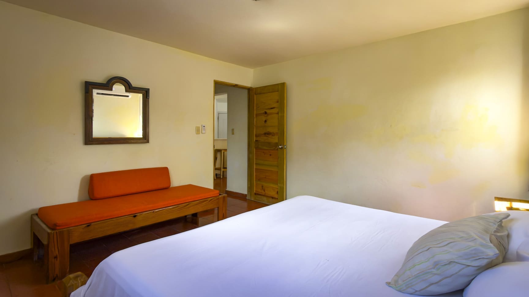 Bright hotel bedroom with white bedding, orange bench, and wooden mirror