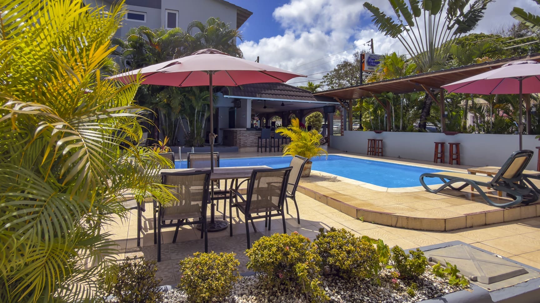 Resort pool with lounge chairs, umbrellas, and tropical plants in sunshine.