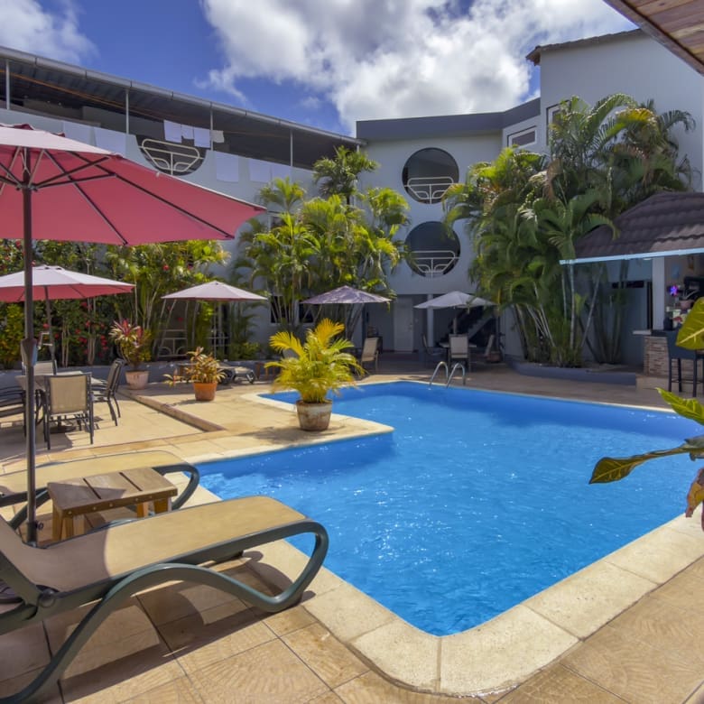 Sunny hotel pool area with pink umbrellas, lounge chairs, and tropical plants around a blue rectangular pool.