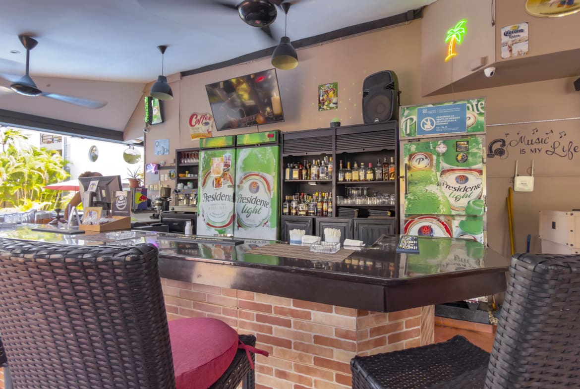 Bar counter with refrigerators, bottles, and bar stools in a tropical setting.