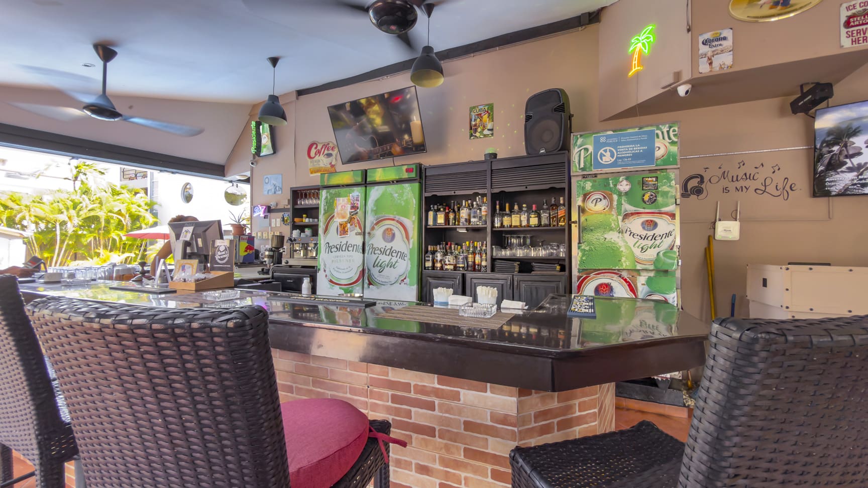Bar counter with refrigerators, bottles, and bar stools in a tropical setting.