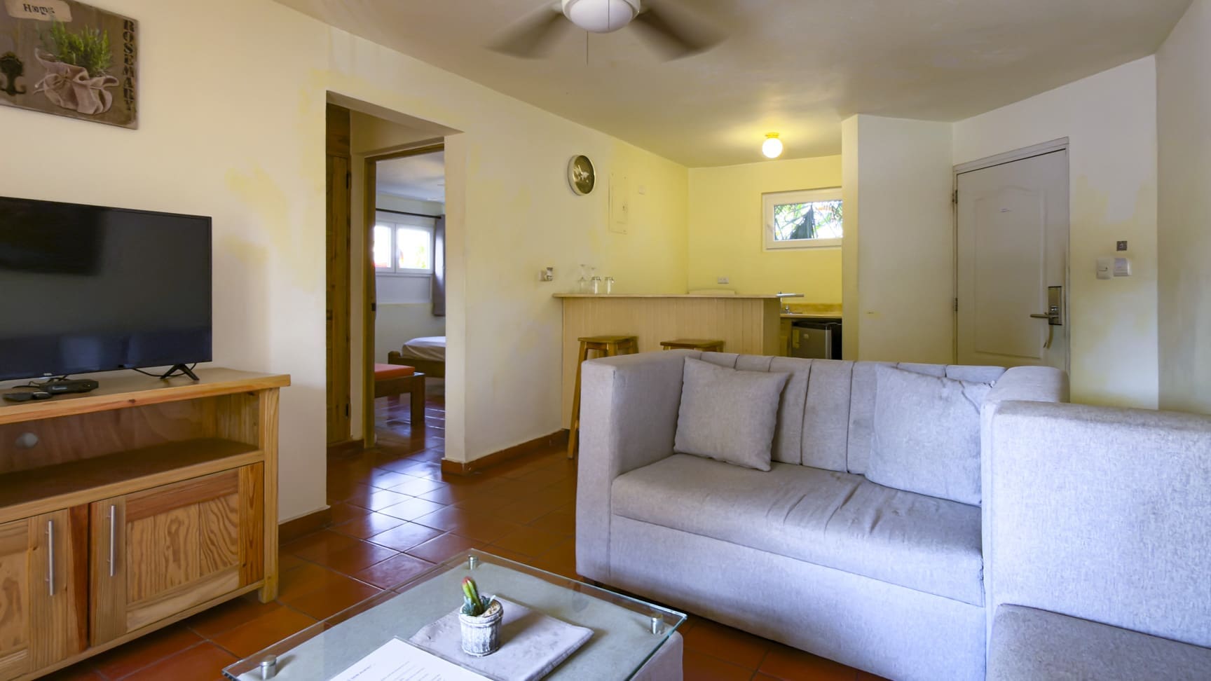 Living room with sofa, TV, and kitchen counter, opening to a bedroom.