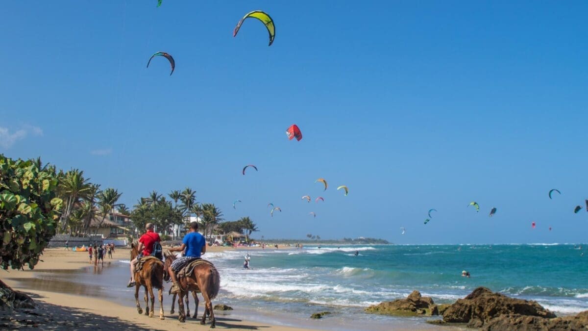 buyDRproperty Two people horseback riding on a beach with kite surfers in the ocean in Cabarete.