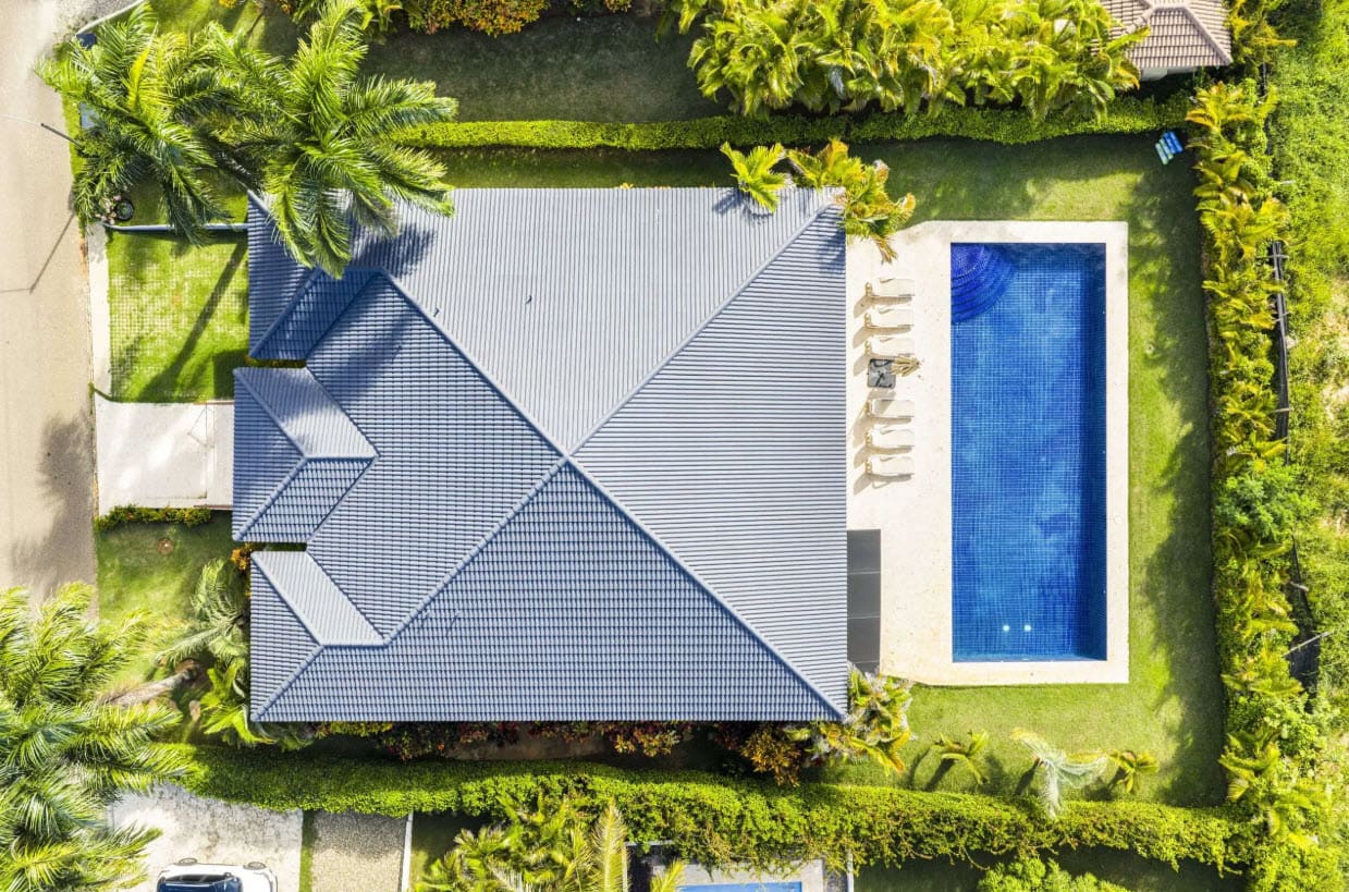 Aerial view of a blue-tiled house with a rectangular blue swimming pool in a fenced yard, surrounded by green lawns and palm trees.