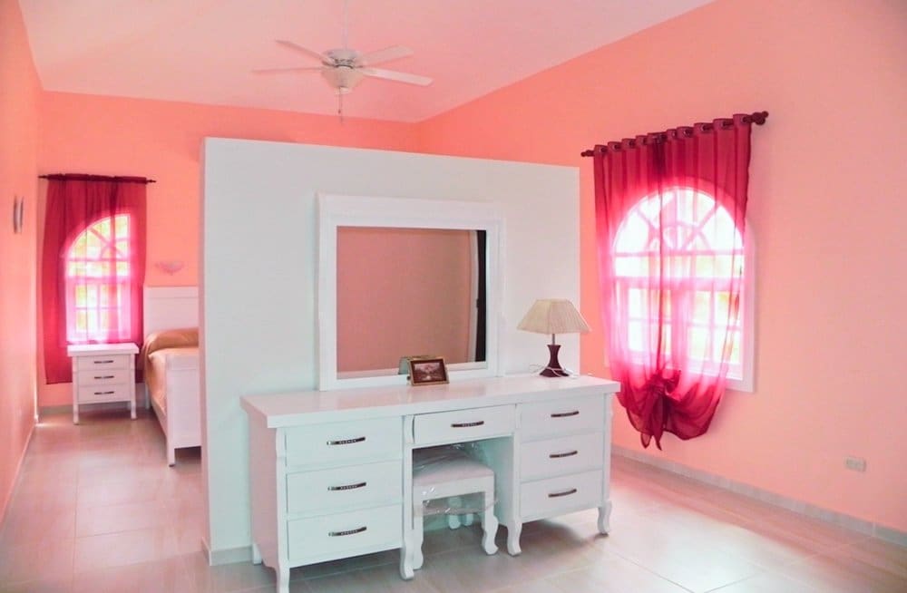 buyDRproperty Pink bedroom with white dresser, vanity stool, and arched windows with red curtains. Sosua Real Estate