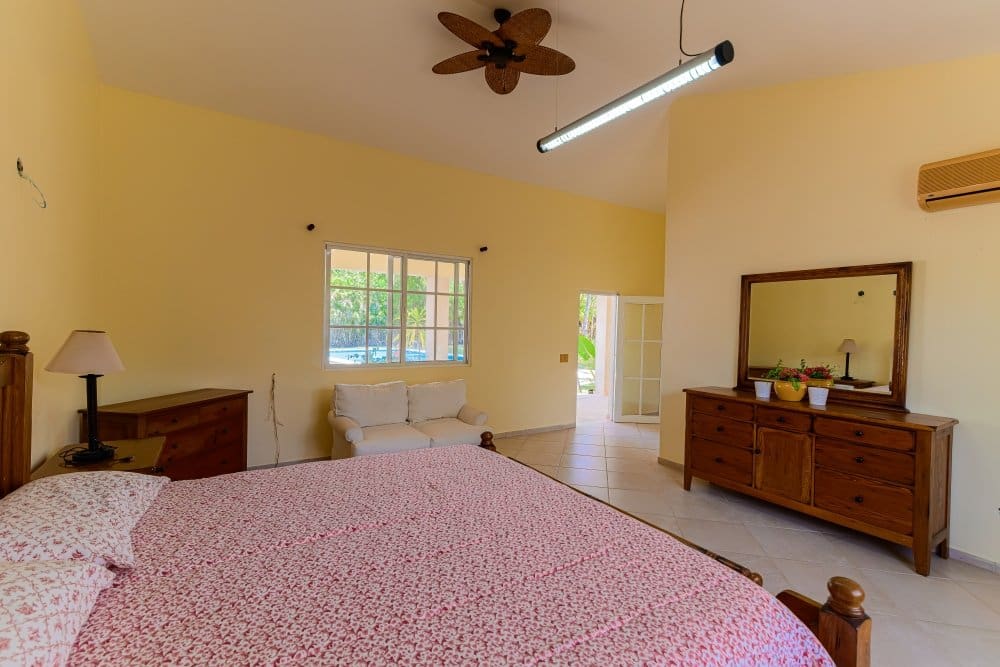 buyDRproperty Yellow bedroom with a large bed, dresser, and window overlooking a pool Sosua Real Estate
