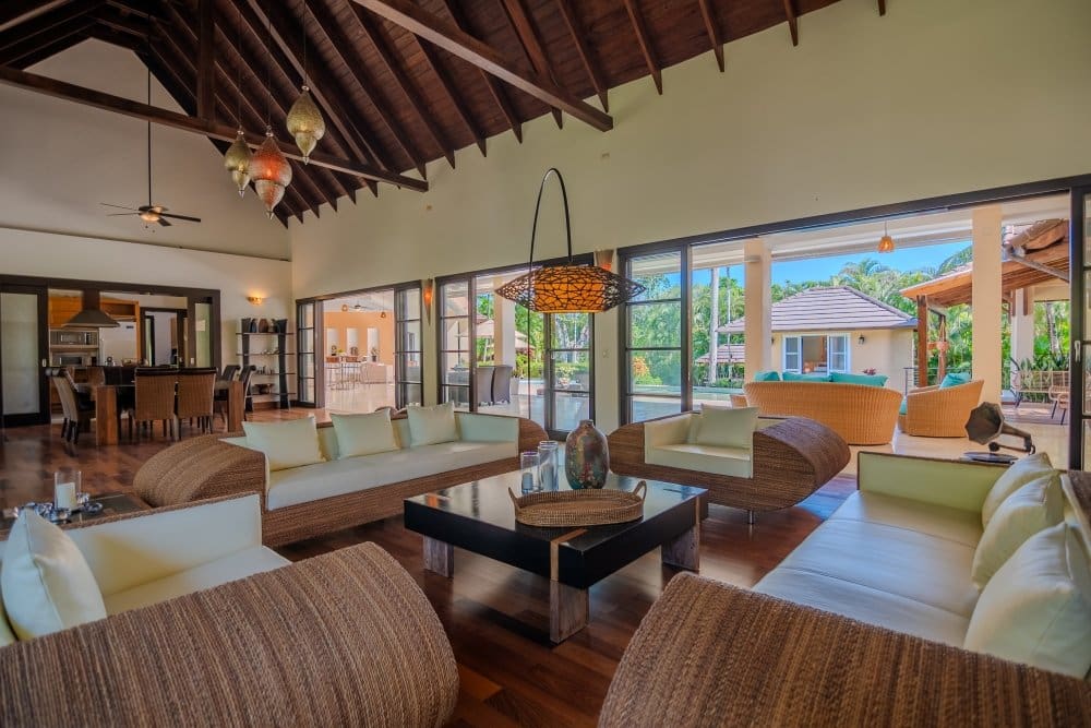 Luxurious open-plan living room with rattan sofas and white cushions, glass coffee table, and vaulted wooden ceiling opening to a tropical patio.