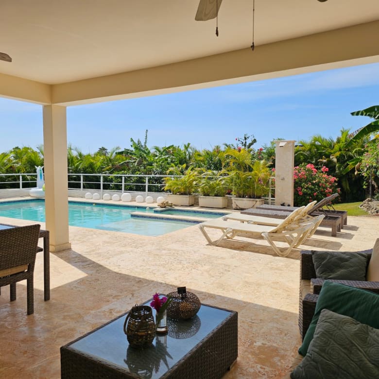 Covered patio area with wicker seating and a glass table overlooking a rectangular pool and tropical garden.