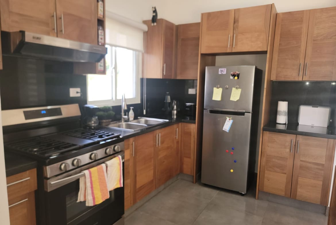 Bright kitchen with stainless steel fridge and a gas range, wooden cabinets, and a dark countertop. A window above the sink lets in light; towels hang from the oven handle.