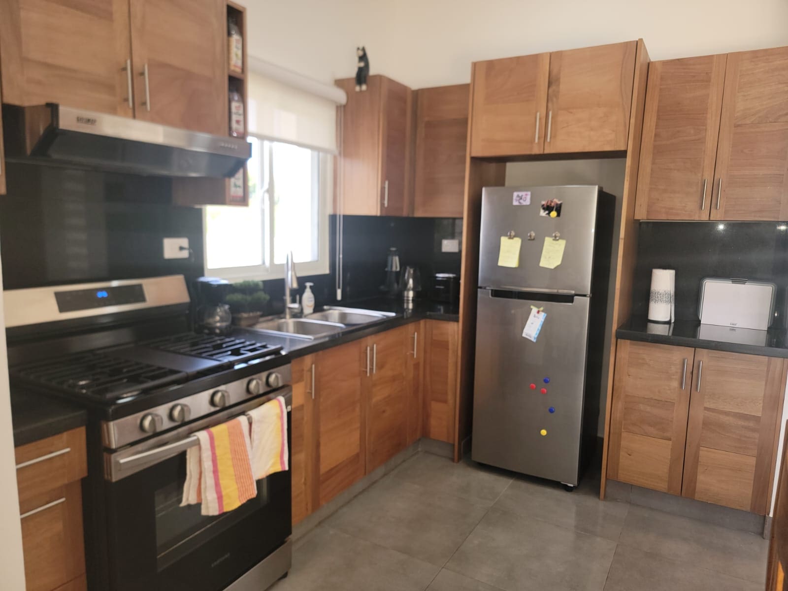 Bright kitchen with stainless steel fridge and a gas range, wooden cabinets, and a dark countertop. A window above the sink lets in light; towels hang from the oven handle.