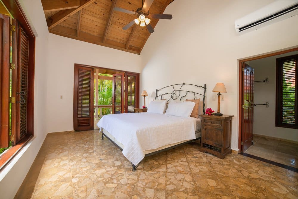 buyDRproperty Tropical bedroom with white bedding, wooden shutters, and AC unit. Sosua Real Estate