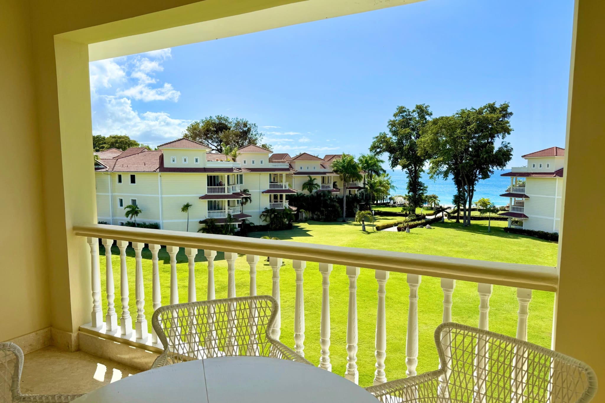 Balcony view of tropical resort buildings, green lawn, and ocean on a sunny day.