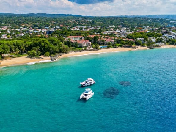 Aerial view of a tropical beach with two yachts in turquoise water and lush green hills.