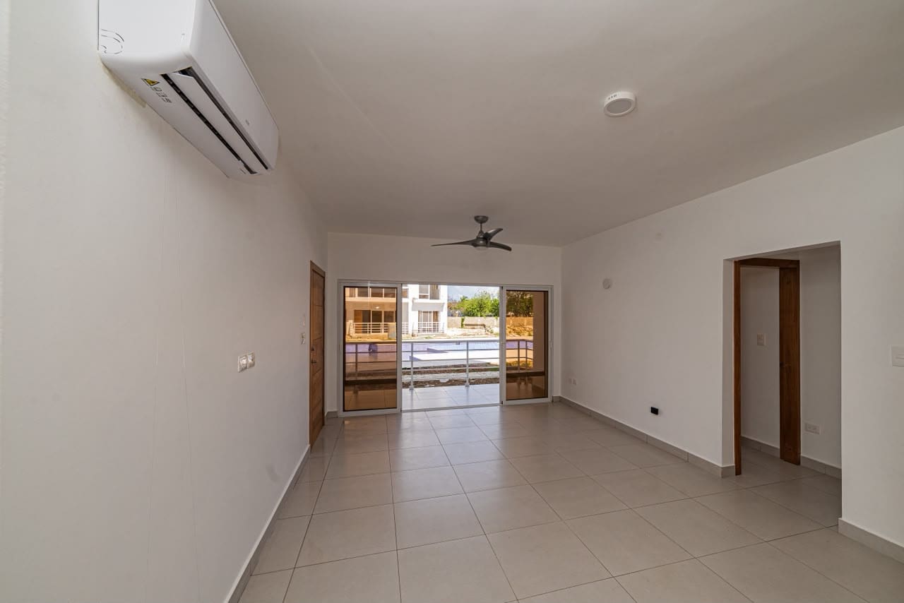 Bright, empty living room with tiled floor, a wall-mounted air conditioner, and a ceiling fan; sliding glass doors open to a balcony with a view of a pool area outside.