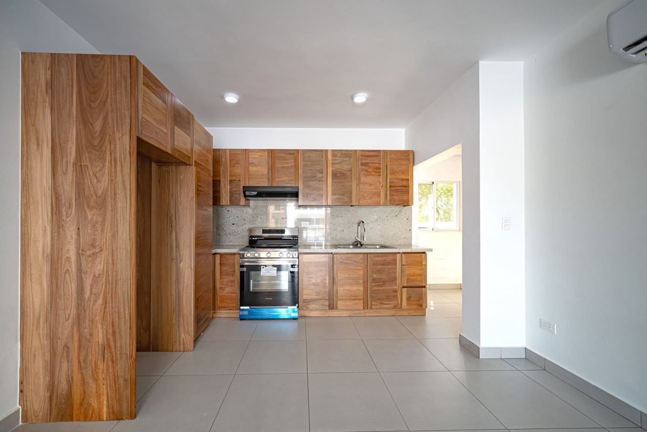 Modern kitchen with warm wood cabinets, stainless steel stove and oven, range hood, and granite countertops. Light gray tiled floor.