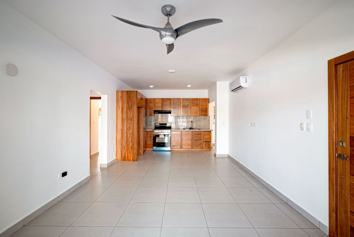 Open-concept living room with tiled floor, white walls, and a wooden kitchen at the back featuring a stainless-steel oven.