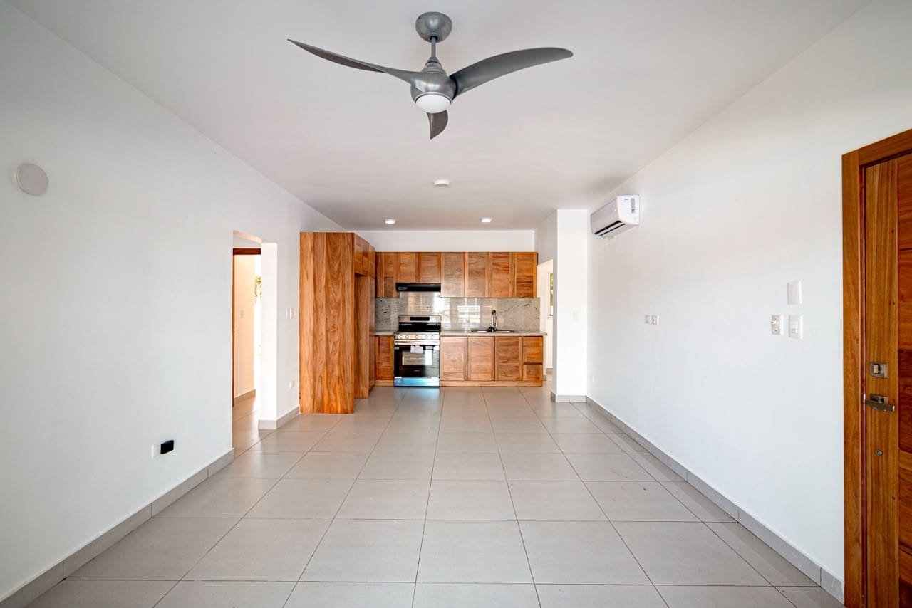 Open-concept living room with tiled floor, white walls, and a wooden kitchen at the back featuring a stainless-steel oven.