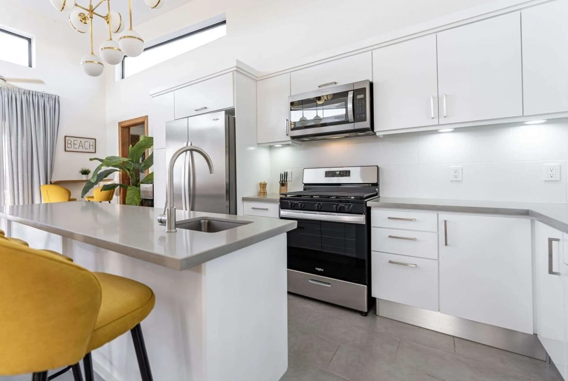 buyDRproperty Modern white kitchen with stainless steel appliances, island, and yellow stools. Sosua Real Estate