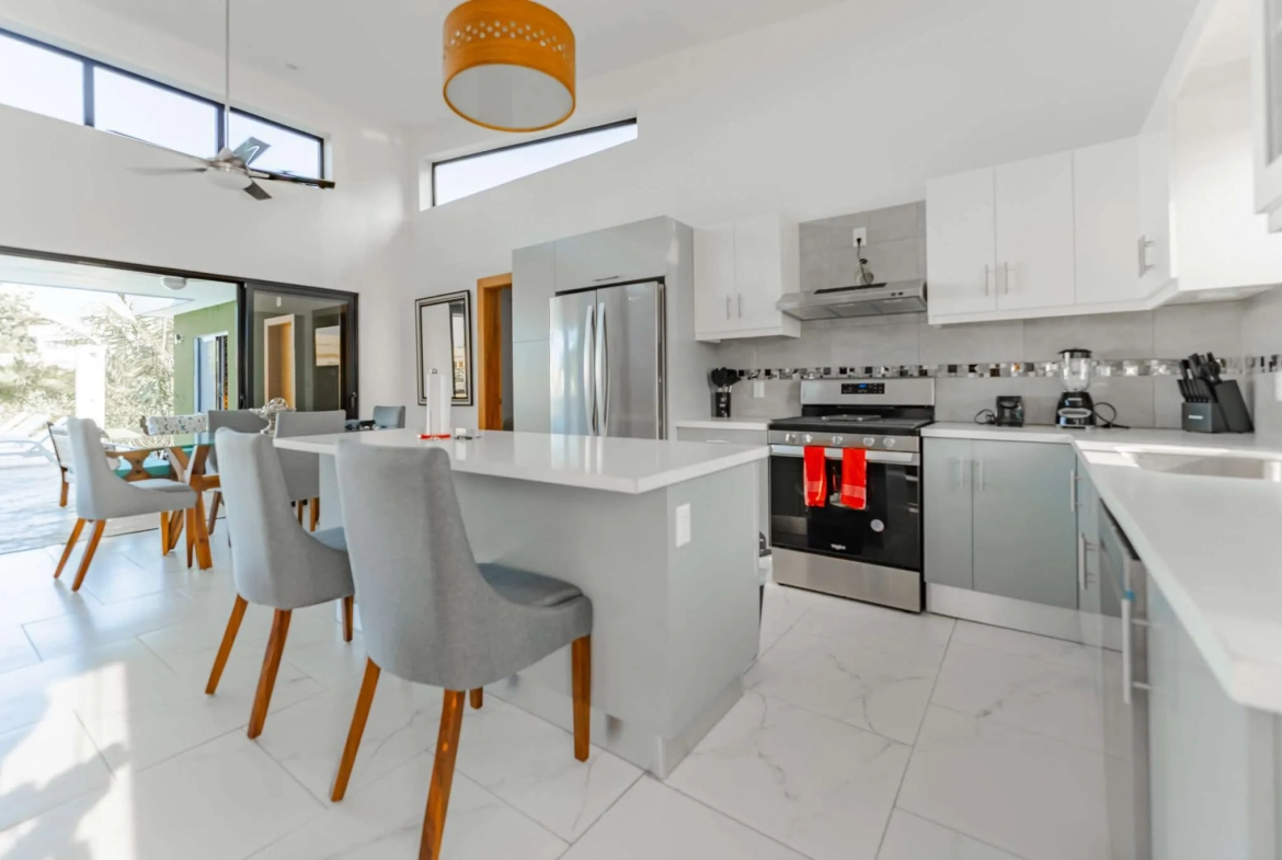 Bright modern kitchen with white cabinets, stainless steel appliances, and a large island with gray stools and wooden legs.