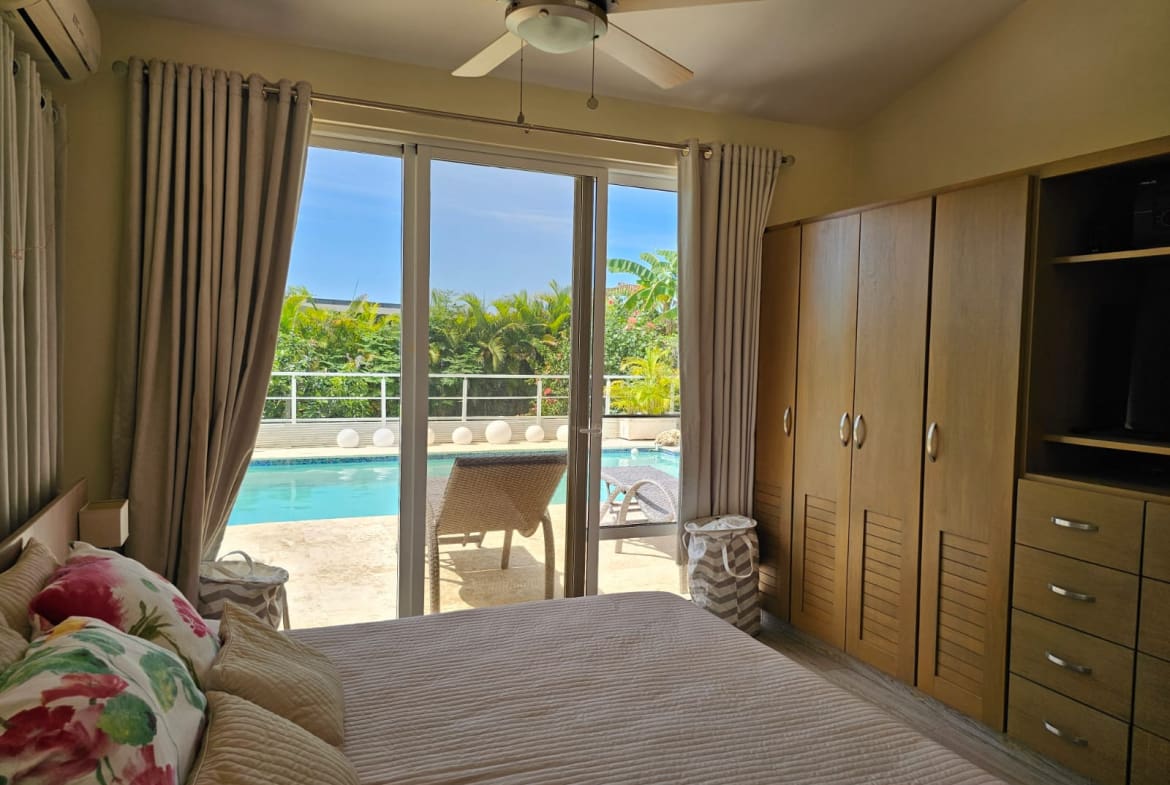 buyDRproperty Bedroom with king-size bed and pool view, tropical foliage, and ceiling fan. Sosua Real Estate