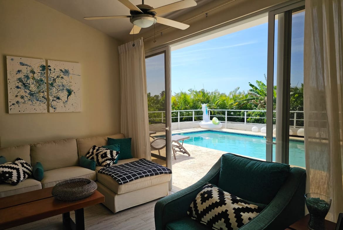 Living room with beige sofa and teal chairs, open to a sunny pool deck through sliding glass doors with white curtains.