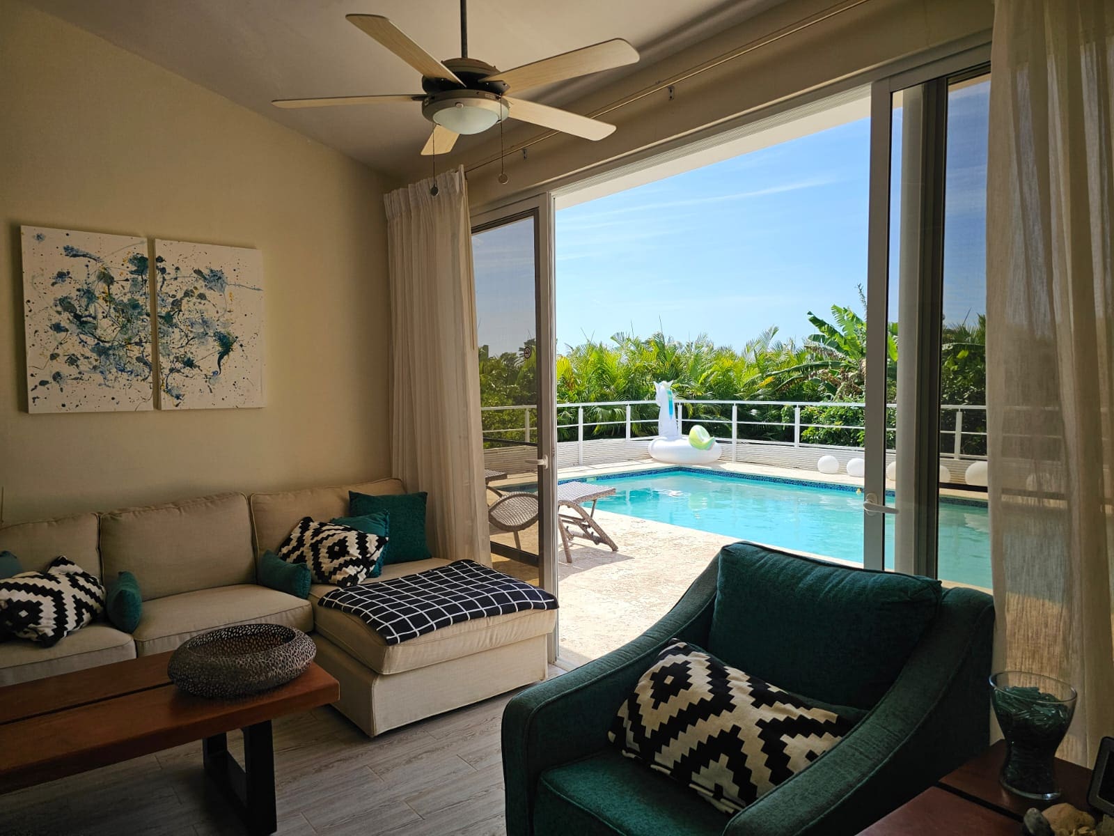 Living room with beige sofa and teal chairs, open to a sunny pool deck through sliding glass doors with white curtains.