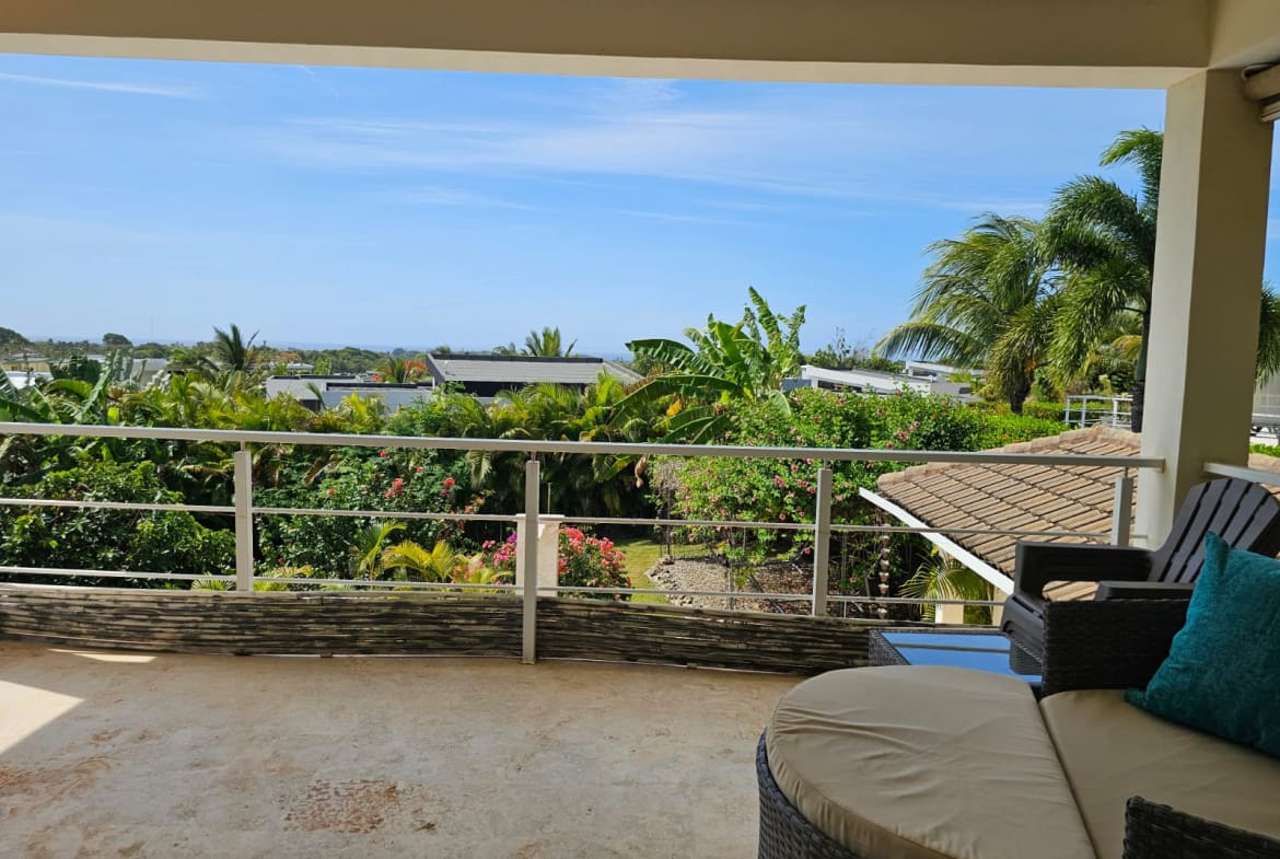 Balcony seating area overlooking a tropical garden with palm trees and distant houses under a blue sky.