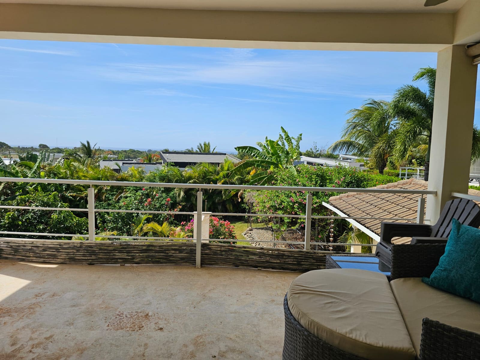 Balcony seating area overlooking a tropical garden with palm trees and distant houses under a blue sky.