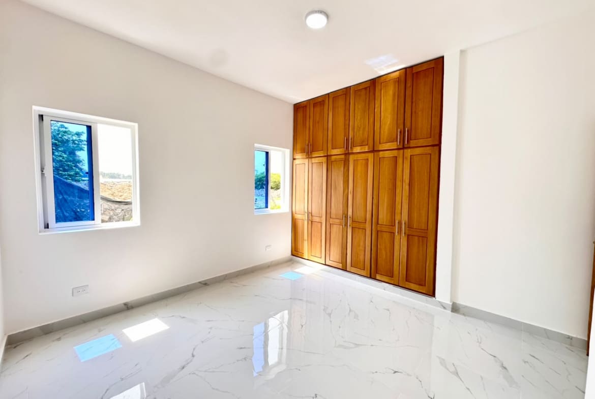 Bright empty bedroom with white walls, glossy marble floor, and tall wooden built-in wardrobes along one wall