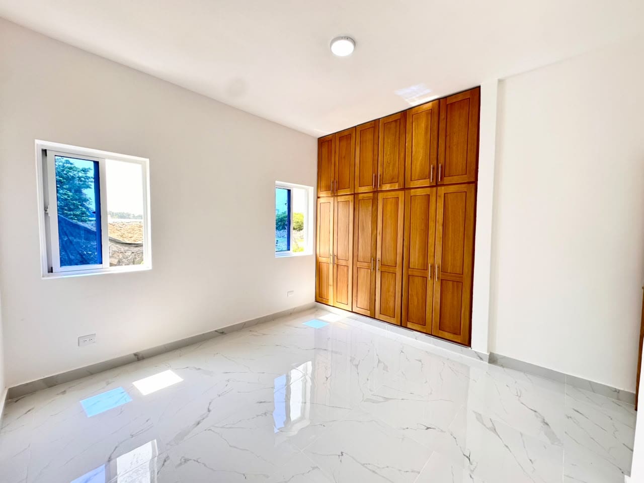 Bright empty bedroom with white walls, glossy marble floor, and tall wooden built-in wardrobes along one wall