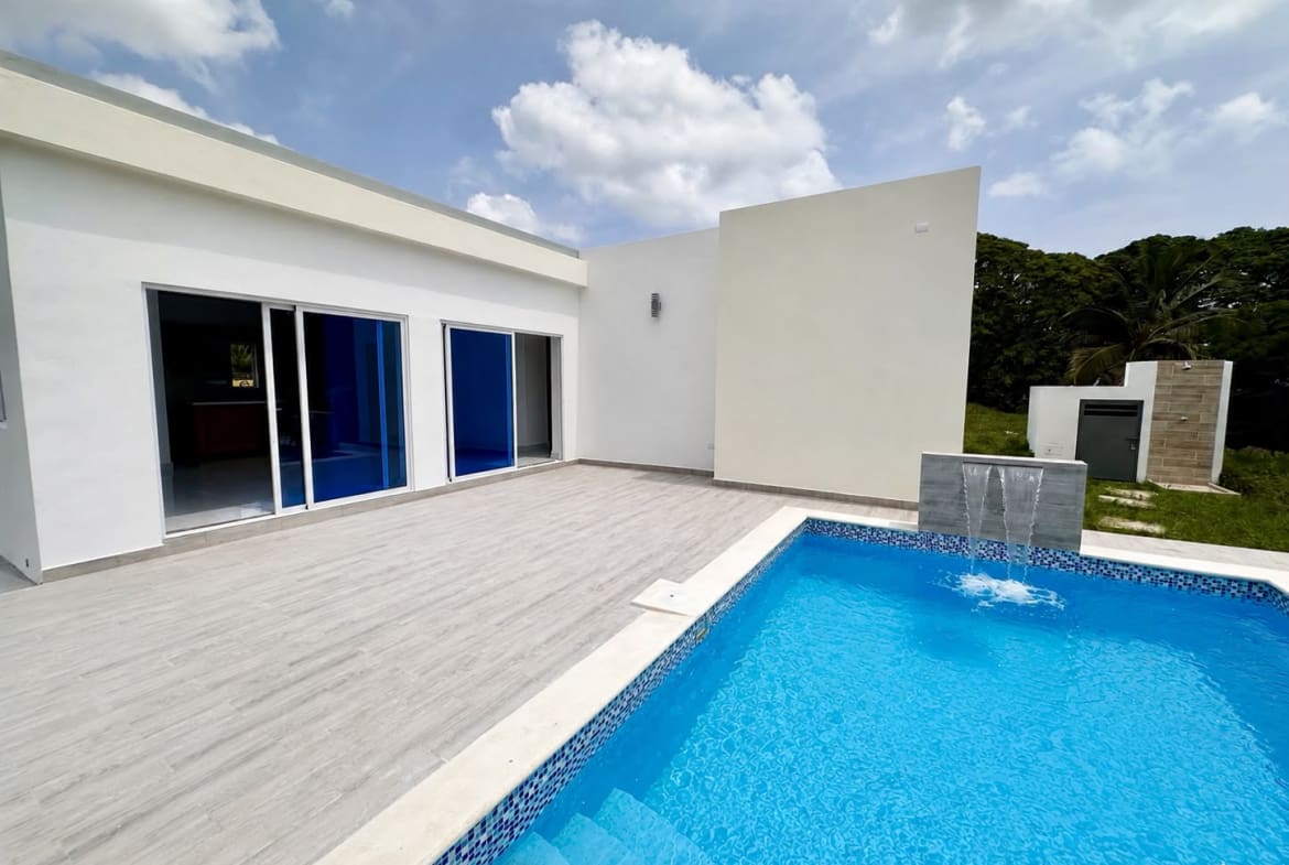 Modern white house with large sliding glass doors opening to a gray tiled patio beside a blue rectangular pool with a small fountain wall at one end, under a partly cloudy sky.