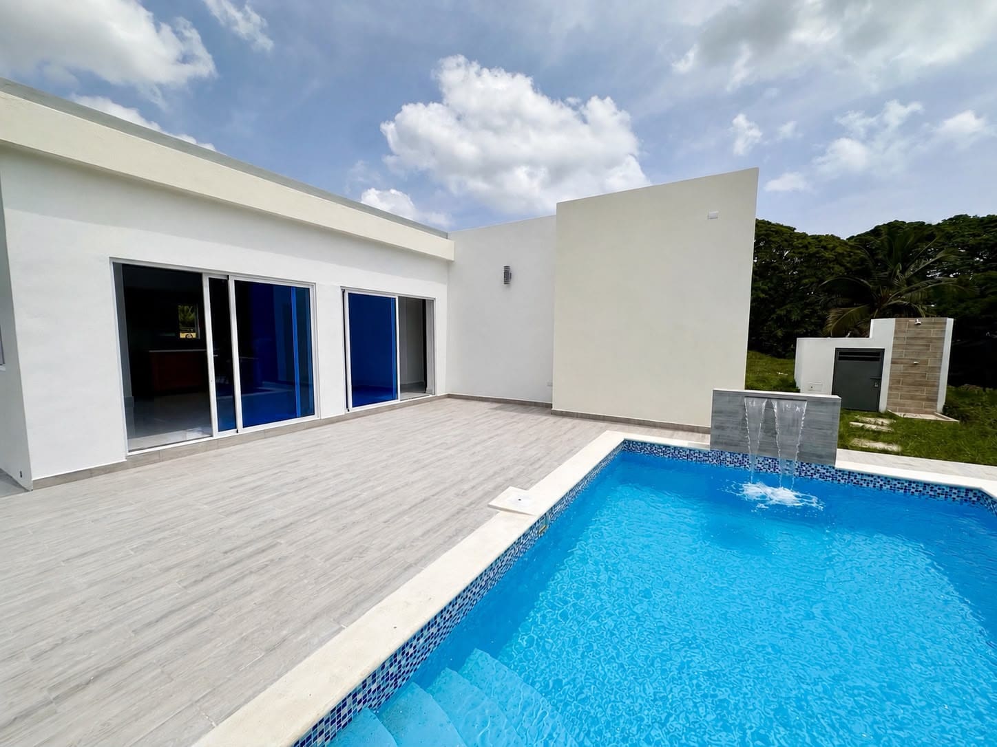 Modern white house with large sliding glass doors opening to a gray tiled patio beside a blue rectangular pool with a small fountain wall at one end, under a partly cloudy sky.