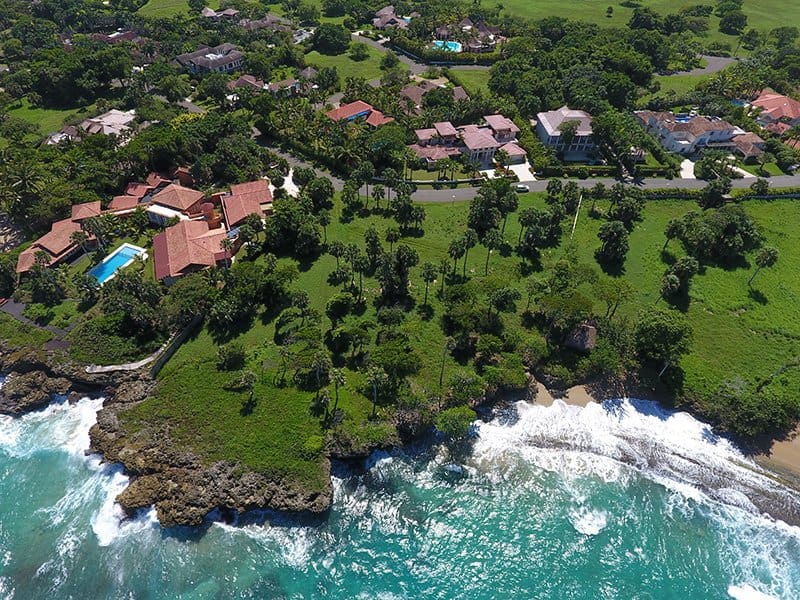 Aerial view of luxury villas and lush green landscape on a tropical coastline with turquoise ocean waves.