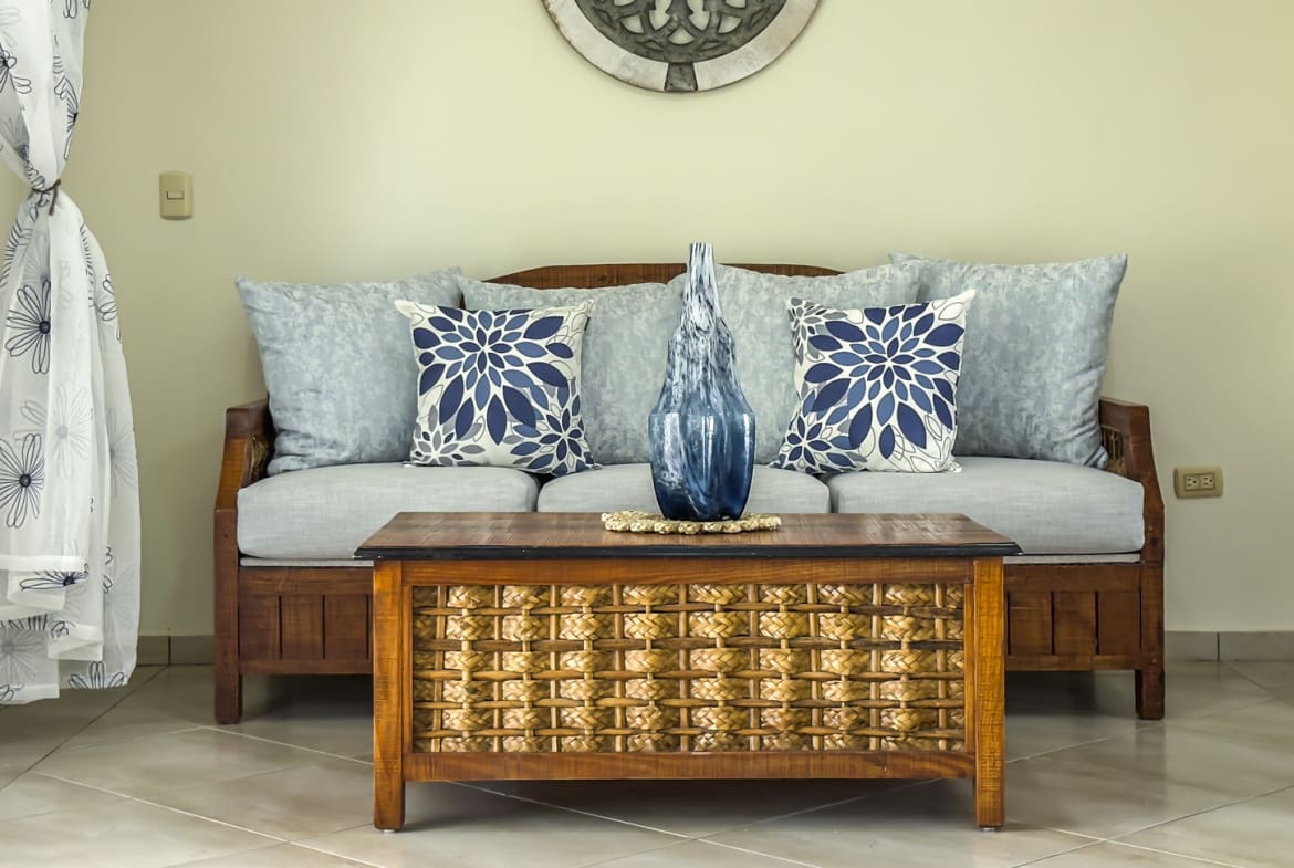 Living room with a wooden sofa, blue floral pillows, and a woven coffee table.