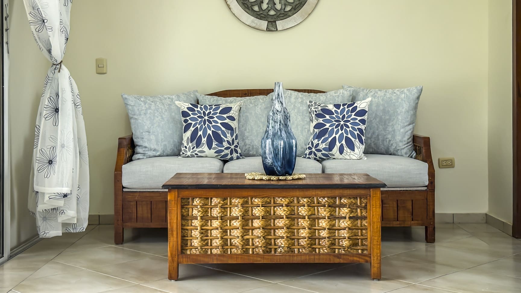 Living room with a wooden sofa, blue floral pillows, and a woven coffee table.