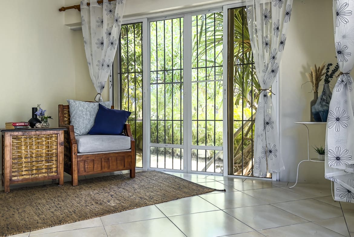 Cozy living room with armchair, woven side table, and floral curtains overlooking lush greenery.