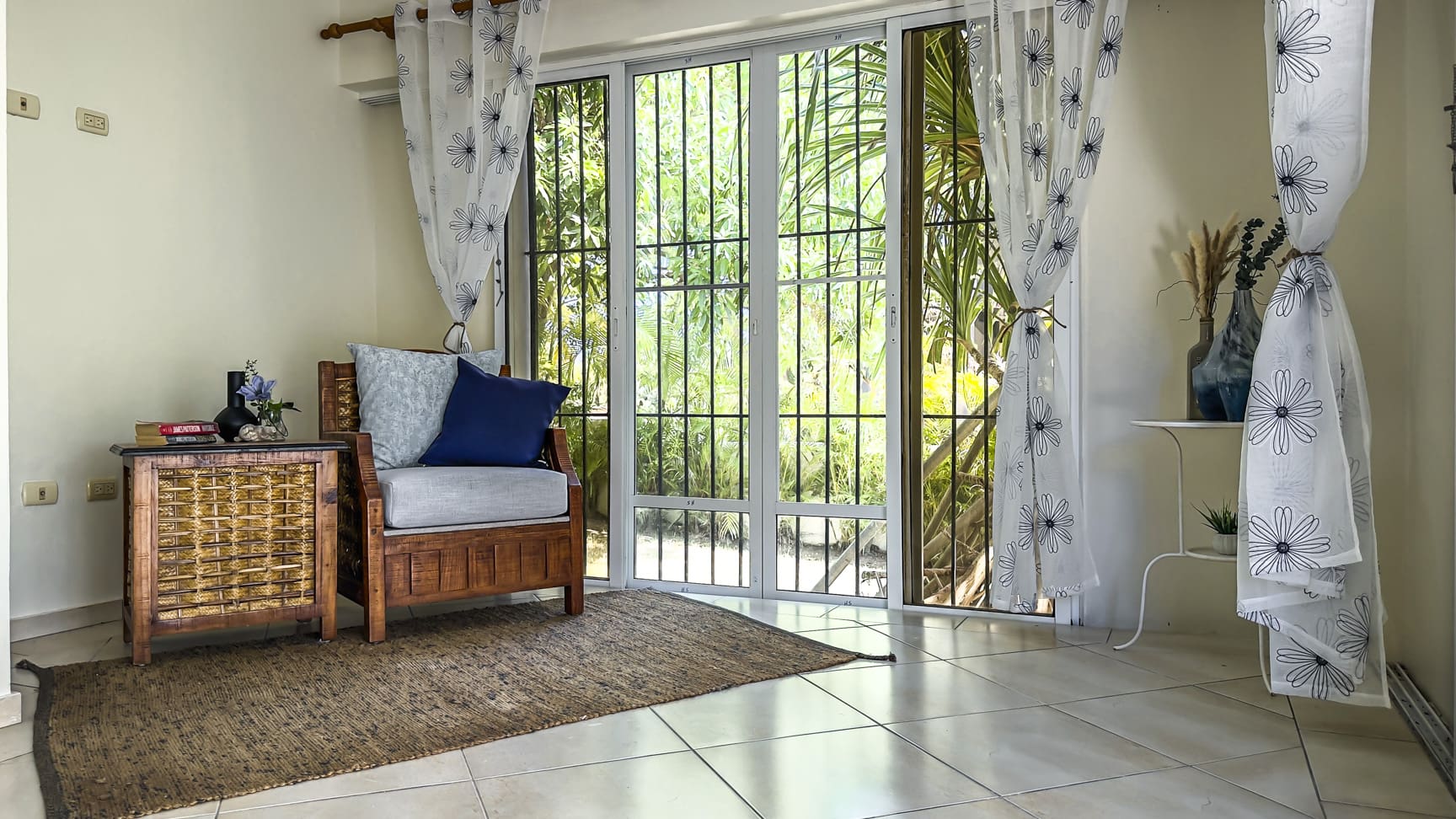 Cozy living room with armchair, woven side table, and floral curtains overlooking lush greenery.