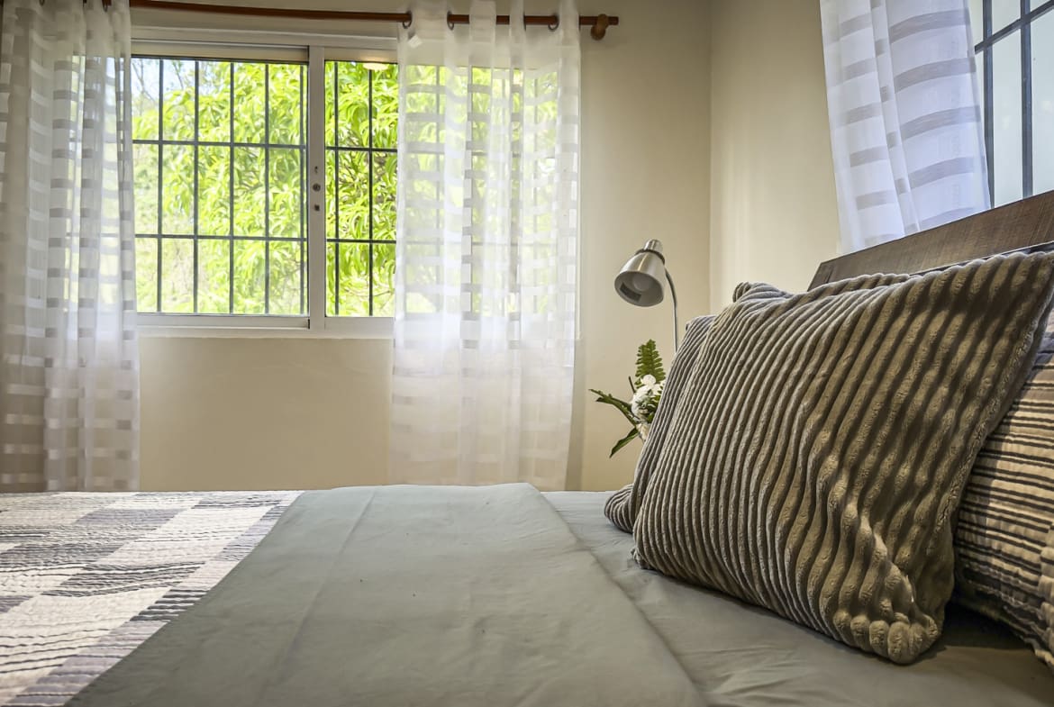 Cozy bedroom with a bed, pillows, and a window with sheer curtains overlooking greenery.