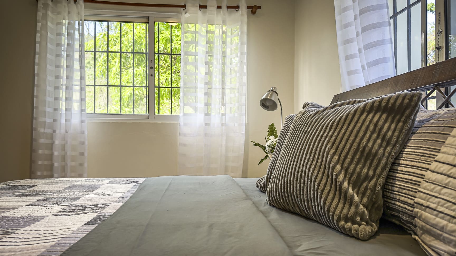 Cozy bedroom with a bed, pillows, and a window with sheer curtains overlooking greenery.