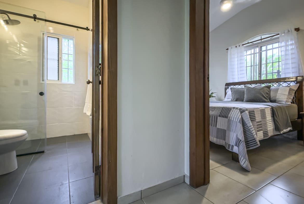 Modern bathroom with white tile and glass shower next to a bedroom with a queen bed.