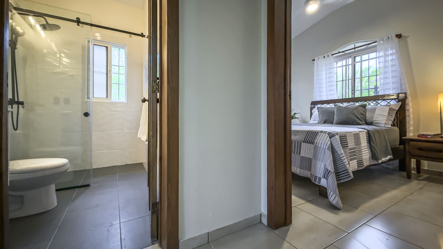 Modern bathroom with white tile and glass shower next to a bedroom with a queen bed.