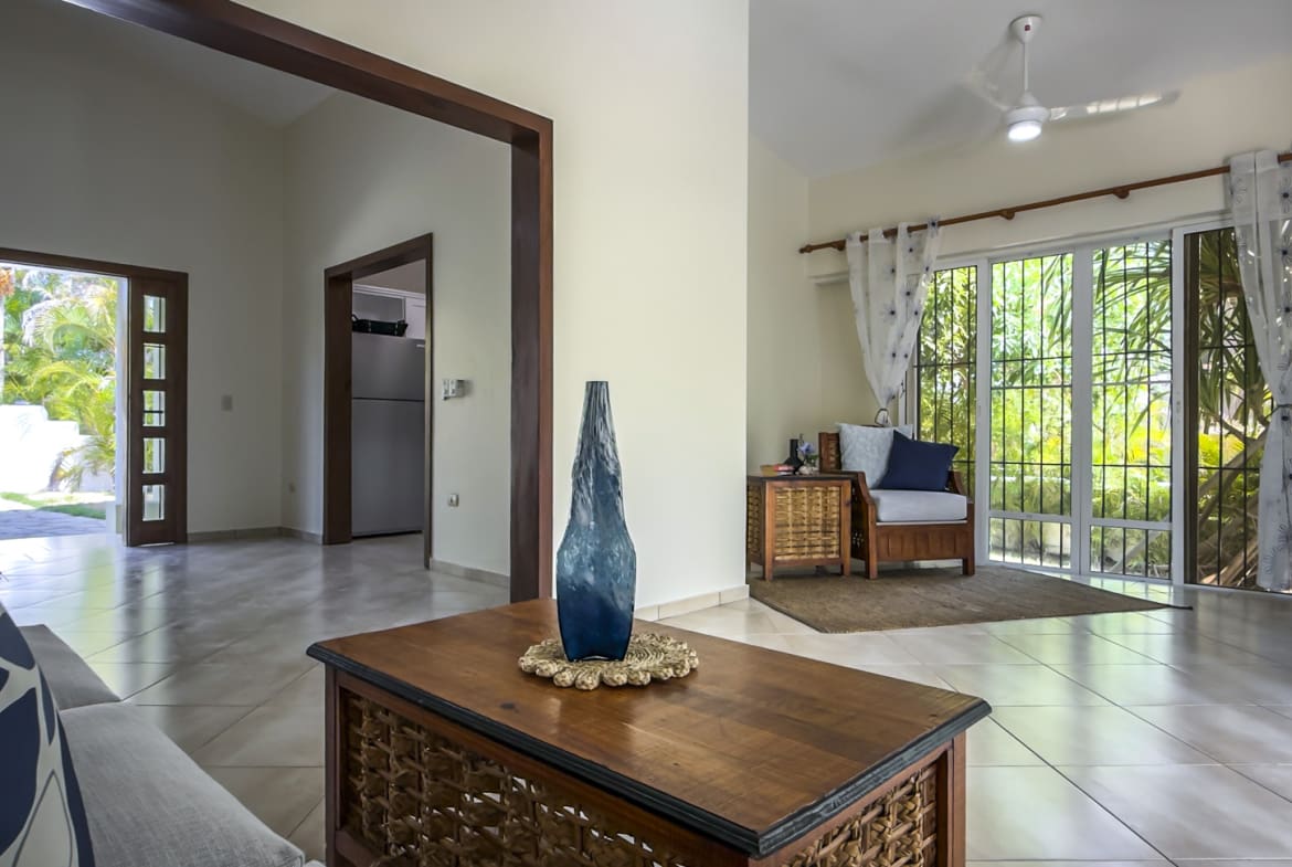 Spacious living room with natural light, featuring a coffee table with a blue vase and a view of a tropical garden.