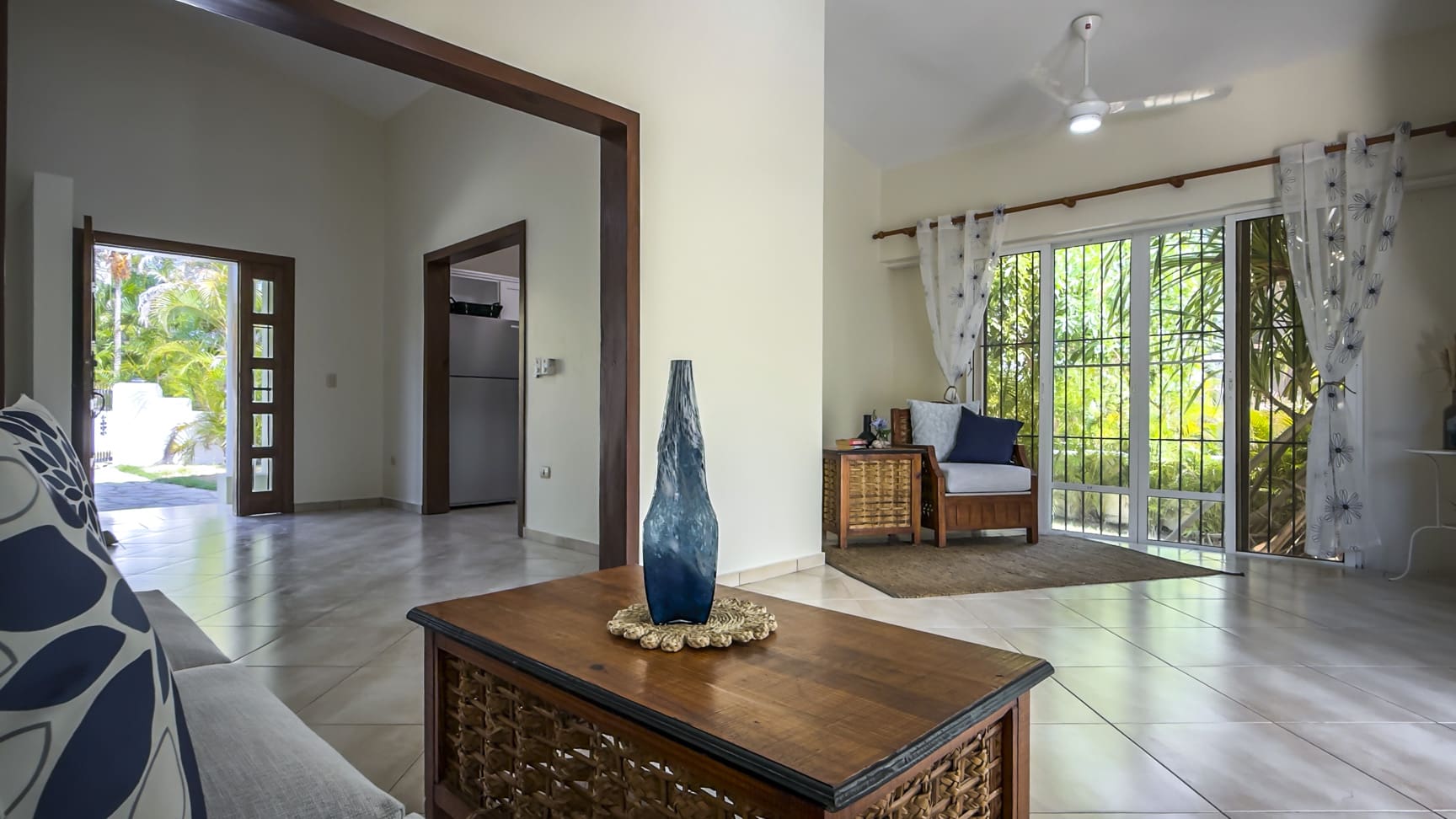 Spacious living room with natural light, featuring a coffee table with a blue vase and a view of a tropical garden.