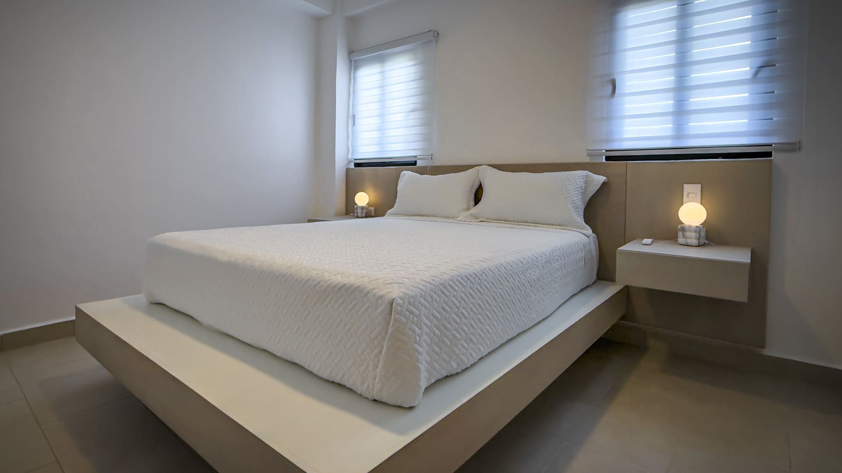 Modern bedroom with minimalist white bed, neutral tones, and ambient bedside lamps.