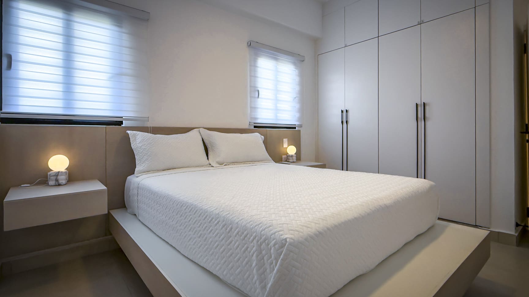 Modern white bedroom with a large bed, minimalist nightstands, and built-in wardrobe.