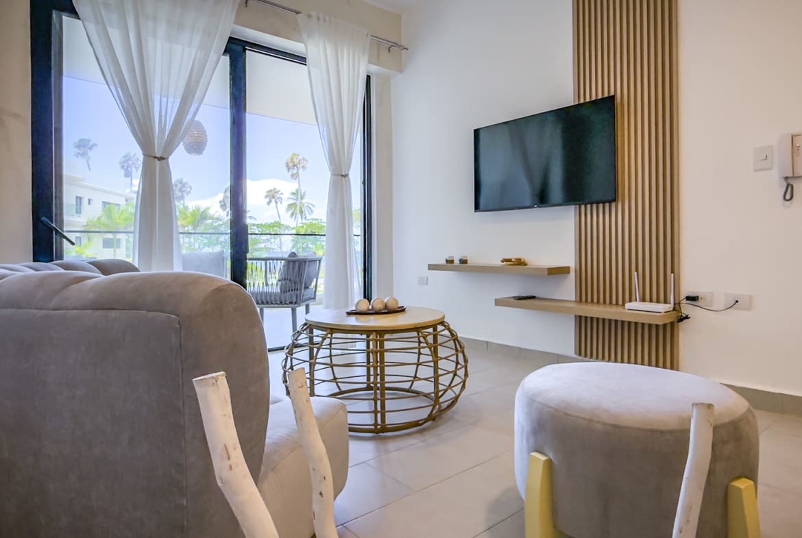 Modern living room with plush sofa, round coffee table, and TV, overlooking a tropical balcony.