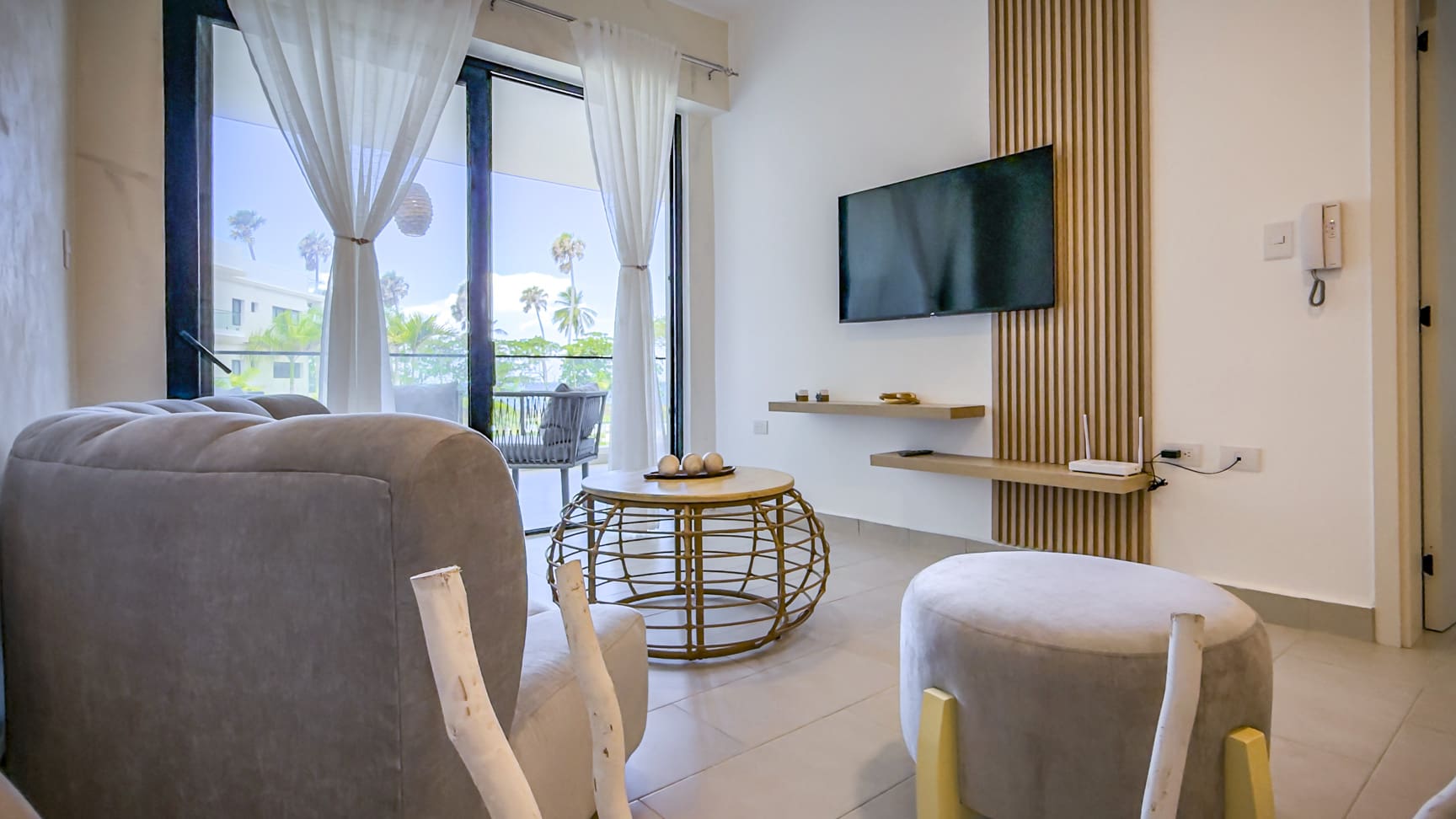 Modern living room with plush sofa, round coffee table, and TV, overlooking a tropical balcony.