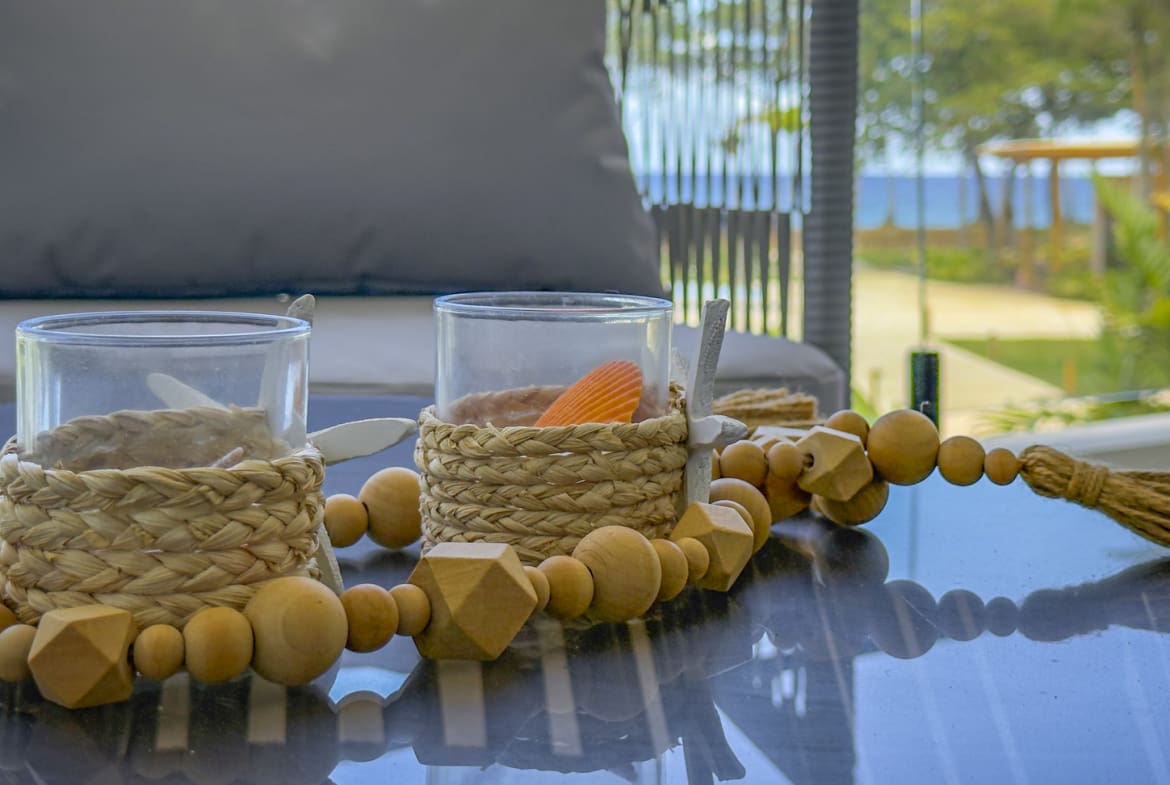 Two glass candle holders with woven natural fiber bases and wooden bead garland on a reflective table.