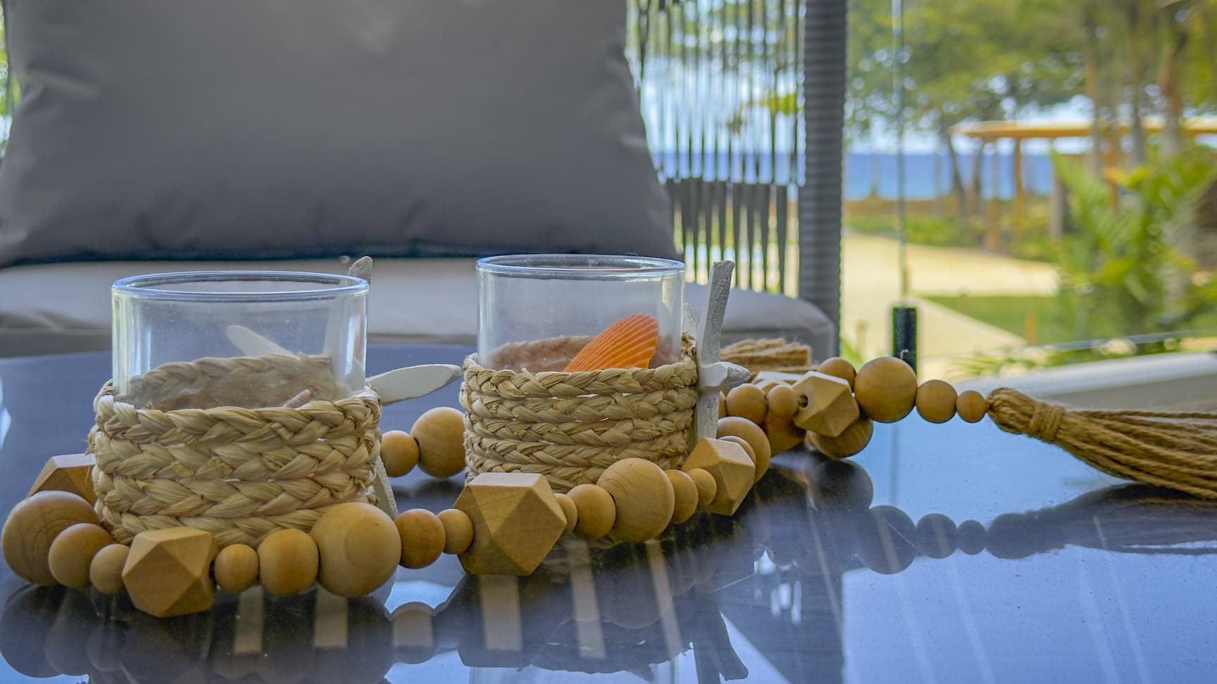 Two glass candle holders with woven natural fiber bases and wooden bead garland on a reflective table.